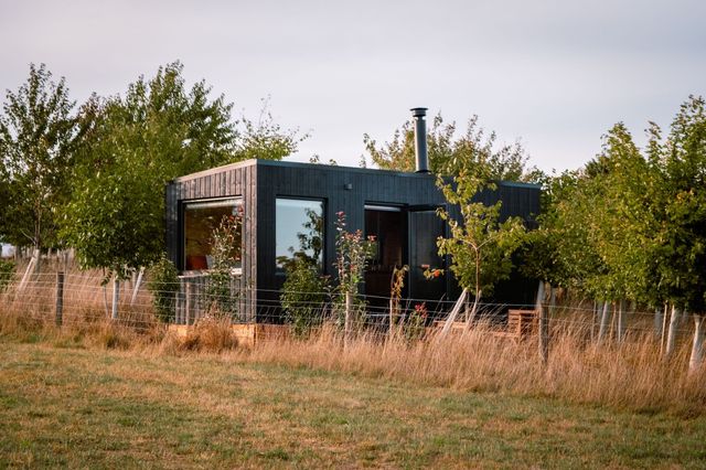 A black timber HIDE n SEEK off-grid cabin among young trees in golden evening light